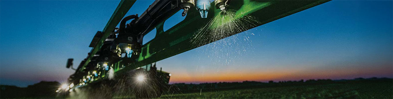 Close-up of a John Deere sprayer boom with active nozzles at dusk, with lights illuminated against a colorful sunset sky.