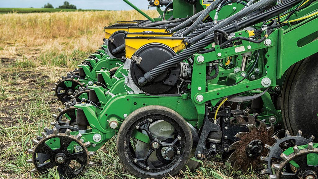 Close-up of John Deere planter row units in a field, showing closing wheels, row cleaners, and seed tubes. Close-up of John Deere planter row units in a field, showing closing wheels, row cleaners, and seed tubes.