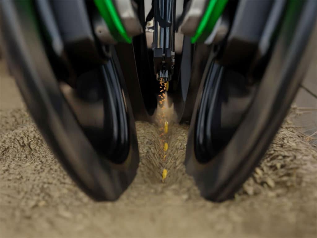 Close-up of a seed drill planting seeds in soil between two black wheels.