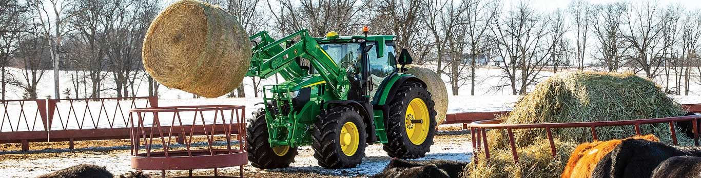 Green tractor lifting a hay bale in snowy field near a feeding ring.