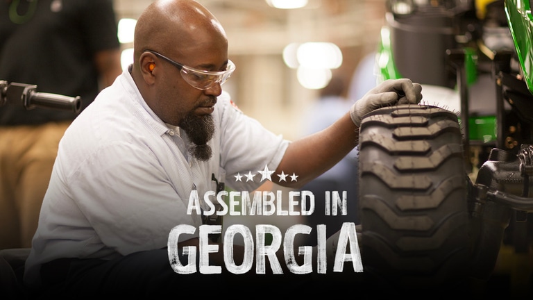 An assembler with eye and ear protection attaches a tire onto a riding lawn mower.