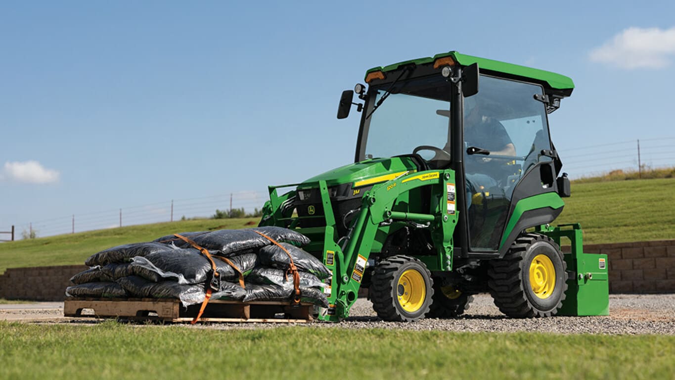 A pallet fork attached to a cabbed green compact tractor lifts a pallet of filled black bags.