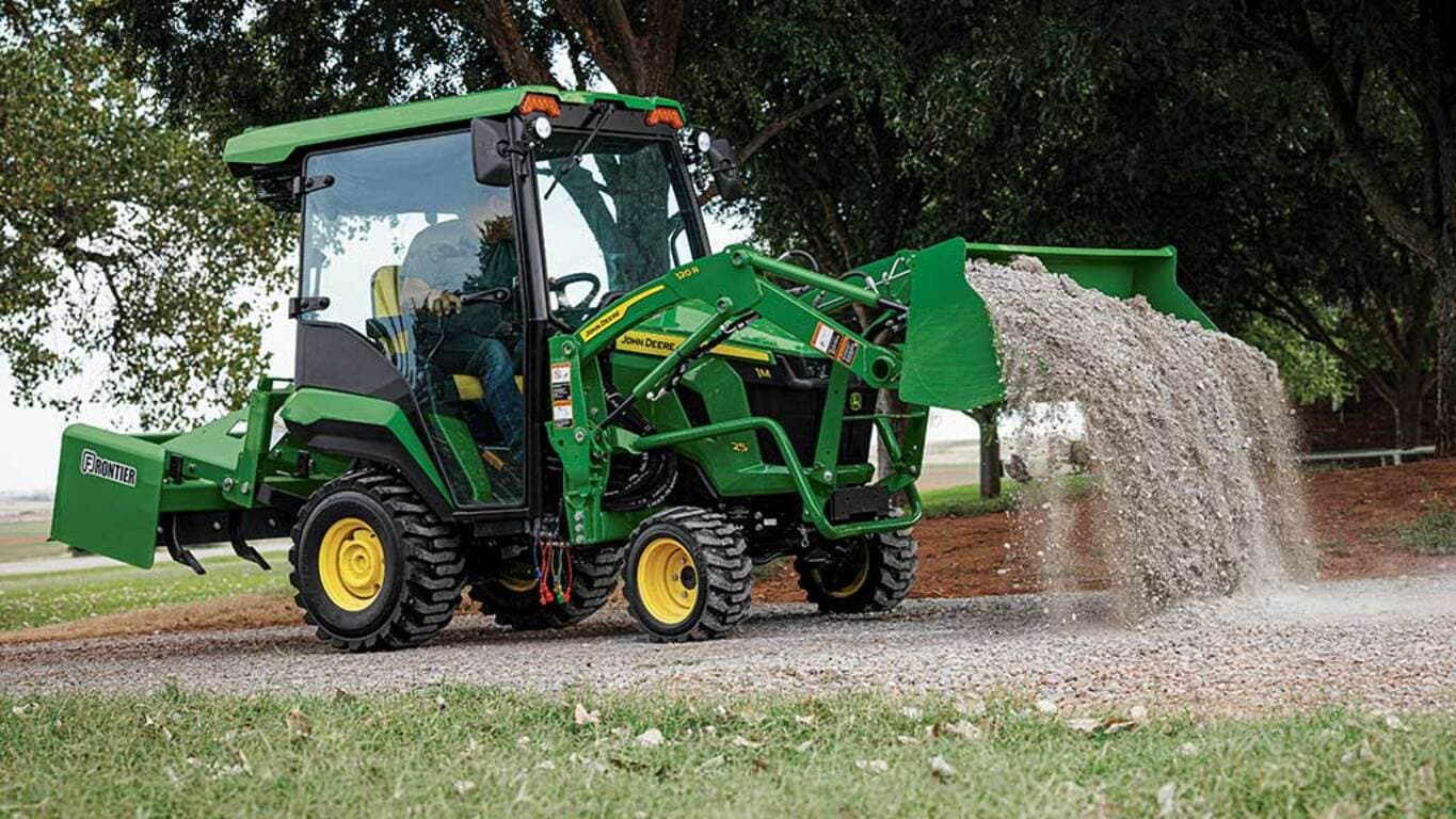 A green compact tractor with a cab, land plane, and loader dumping gravel on a gravel road.