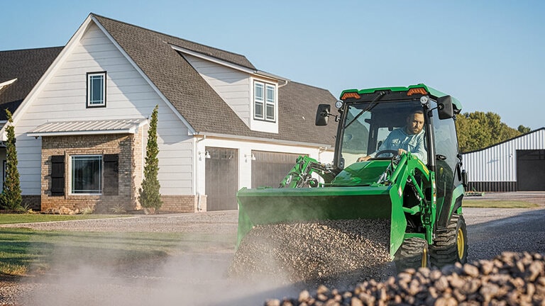 A cabbed green compact tractor using a loader to push gravel down a gravel road.