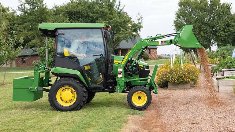 A green compact tractor with a rear ballast box and front loader dumps gravel around some planter boxes.