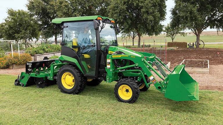 A green compact tractor with a loader and ground seeder drives next to a garden plot.