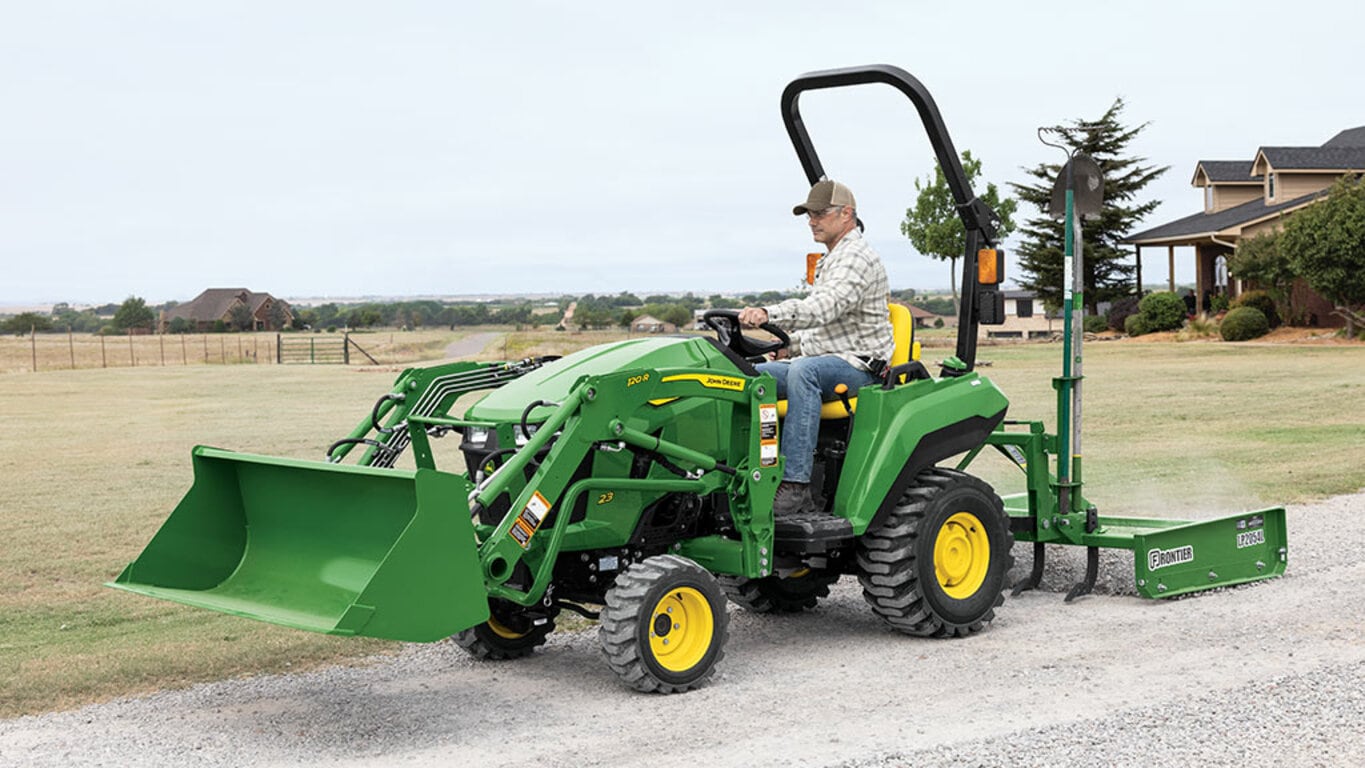 A person driving a green compact tractor equipped with a loader and land plane spreading gravel on a gravel road.