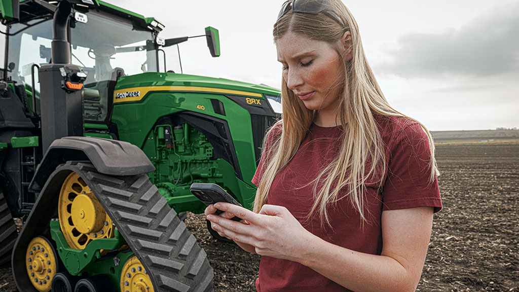 A farmer stands next to John Deere 8RX 410 tracked tractor connecting her phone to her Autonomy Ready tractor