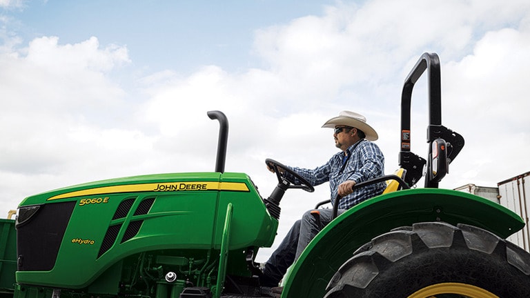 A person in a cowboy hat drives a green tractor with an open operator station.