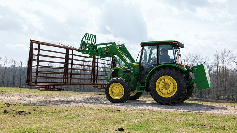 A green tractor uses a pallet fork attachment to carry fence pieces down a gravel road.