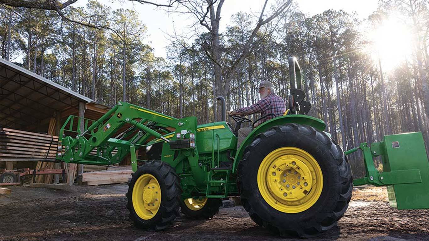A pallet fork attachment is used by a green tractor to move planks under an open storage area.