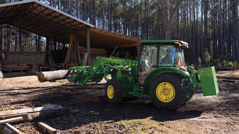 A grapple attachment is used by a green tractor to move logs under an open storage area.