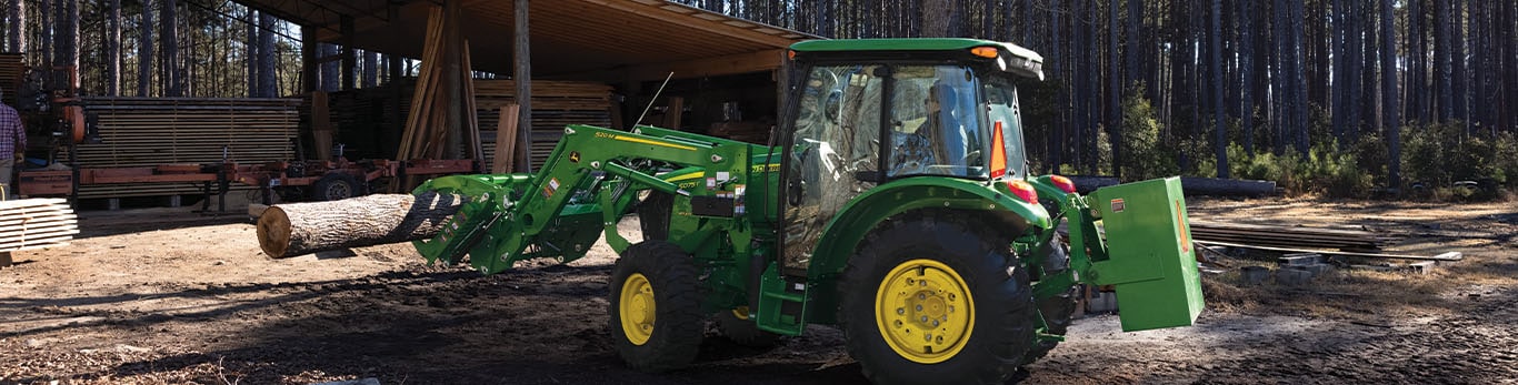 A grapple attachment is used by a green tractor to move logs under an open storage area.
