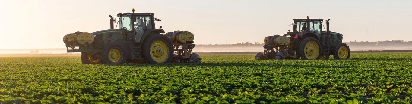 Two tractors equipped with fertilizer tanks work in a lush green field during sunrise.