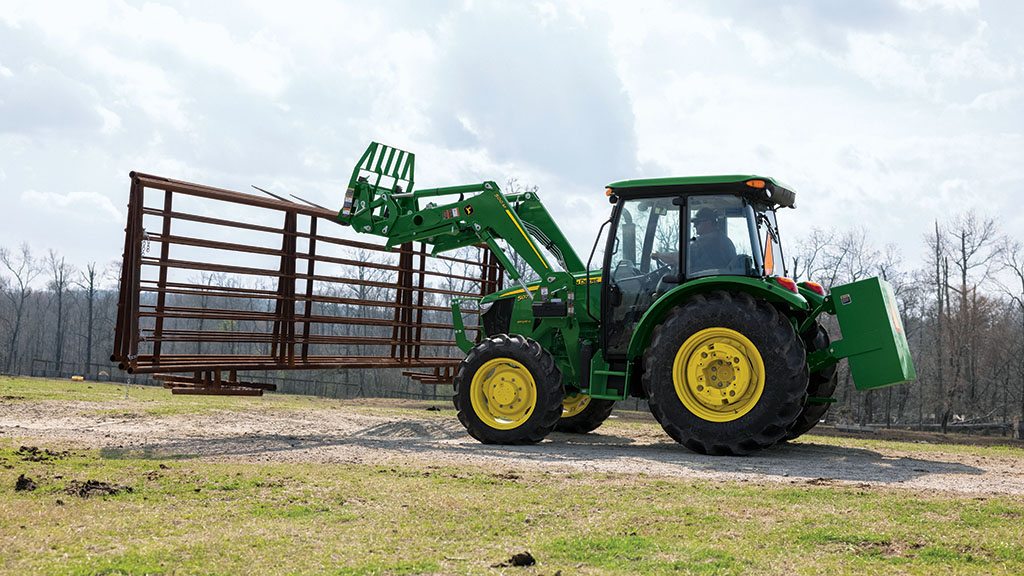 A green and yellow John Deere 5E Series tractor uses a pallet fork attachment to carry fence pieces down a gravel road.