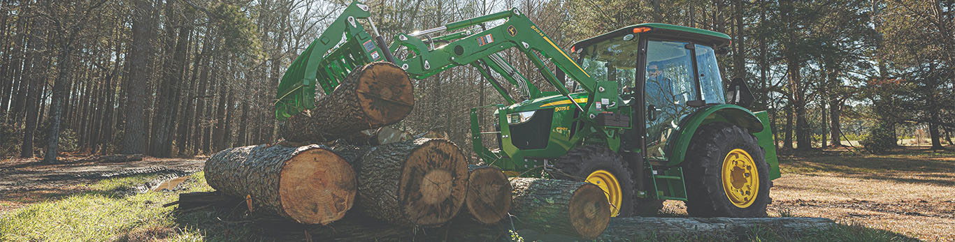 A green tractor with a front grapple attachment lifts large logs in a wooded area.