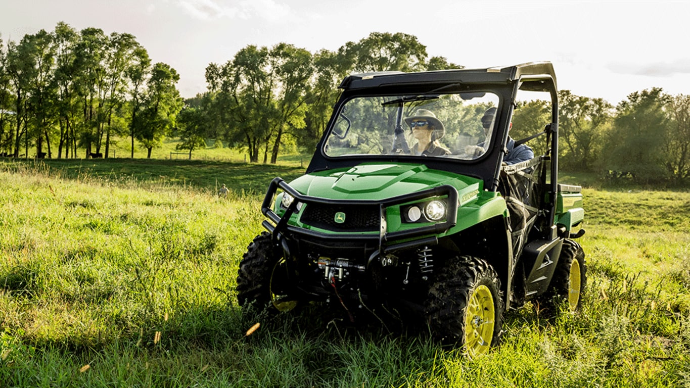 Two people in a green Gator drive in an open grassy field with thin trees in the background.