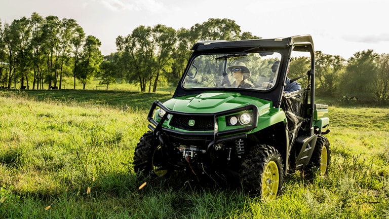 Two people in a green Gator drive in an open grassy field with thin trees in the background.