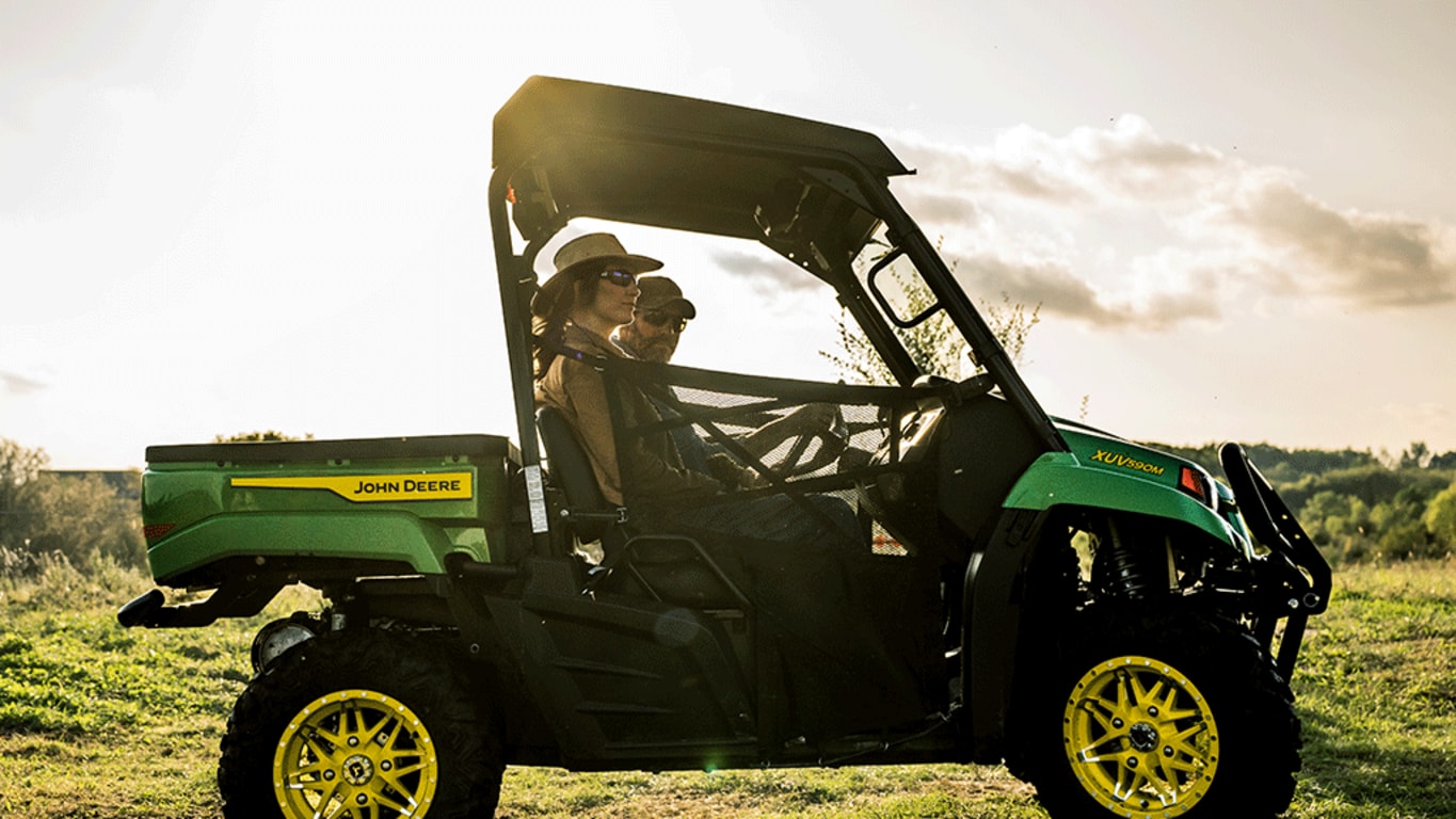 A Gator carrying two passengers off-roads in a field with the sun shining directly behind it.