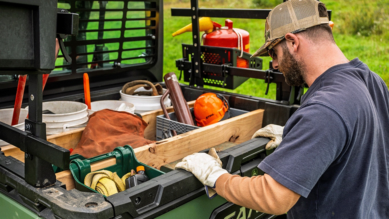 A person looks into a cargo box full of various tools and supplies on the back of an olive-green Gator.