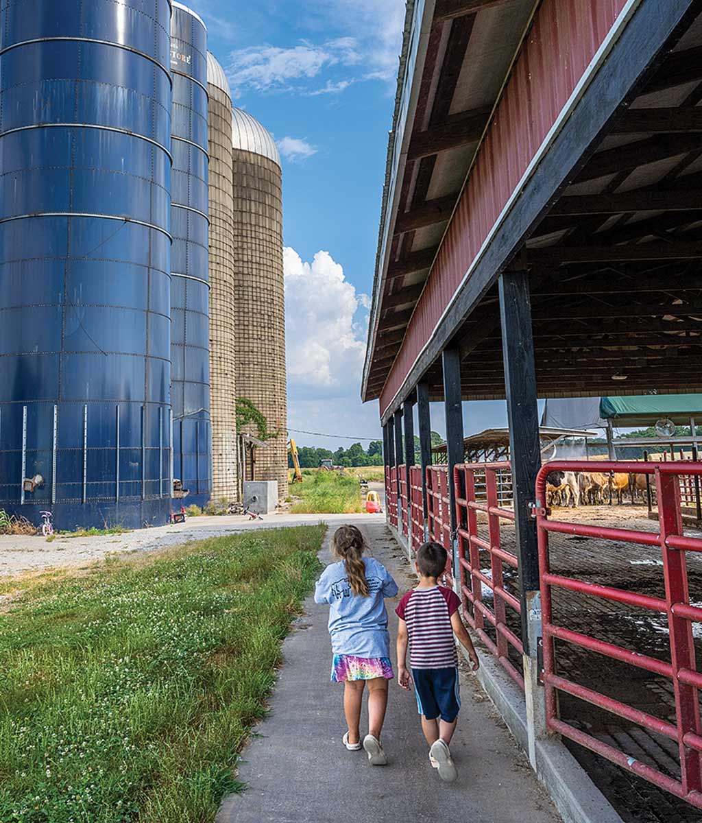 two children walking by cattle pens with silos in distance