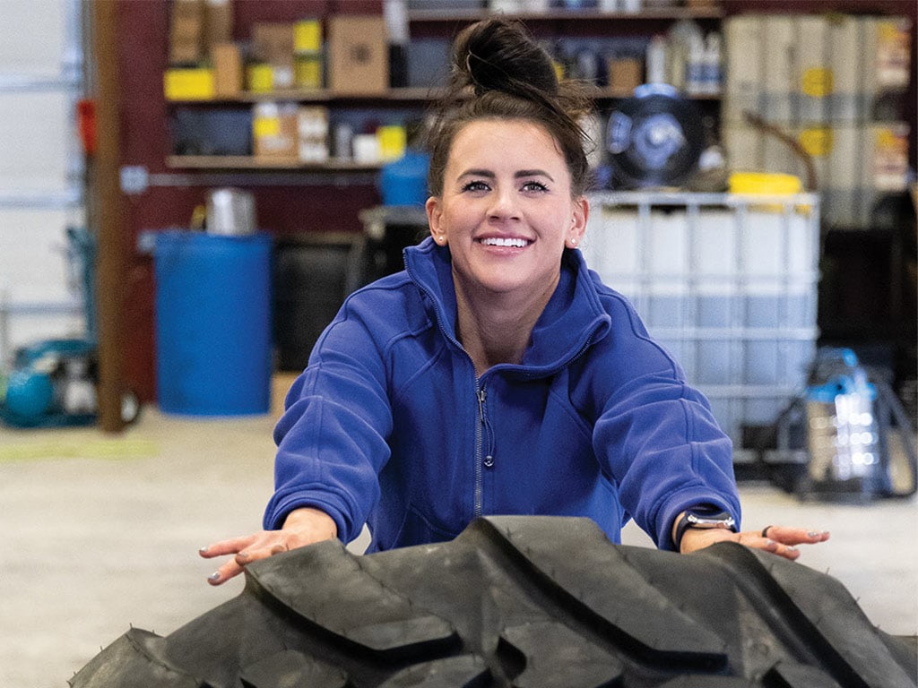 Young woman pushing tractor tire in warehouse with workout sweater on
