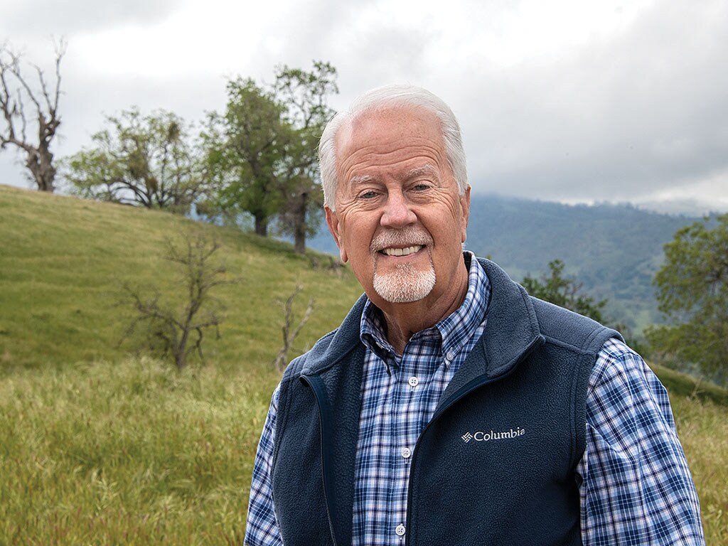 white haired man smiling with hillside and trees in background