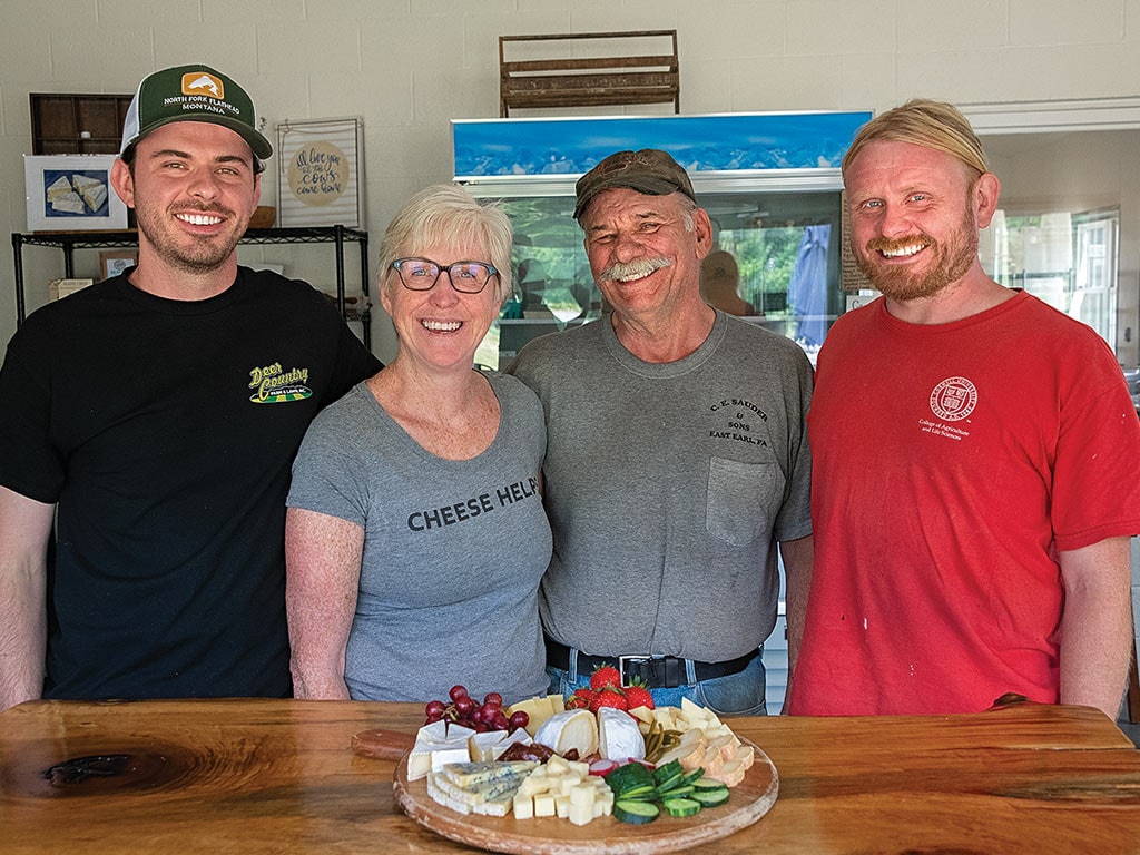 cheesemaker family standing at a wooden table with a round board of cheeses