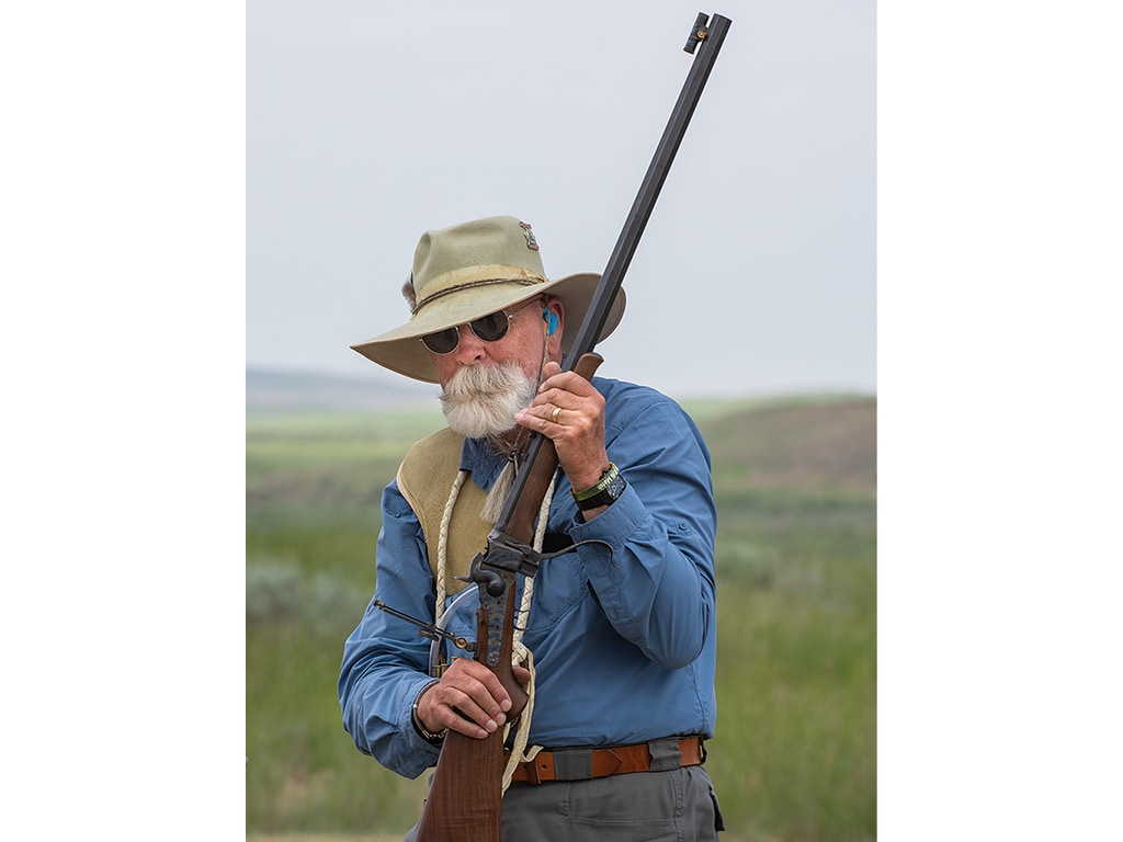  man with white facial hair, sunglasses, and wide brimmed hat holding a rifle