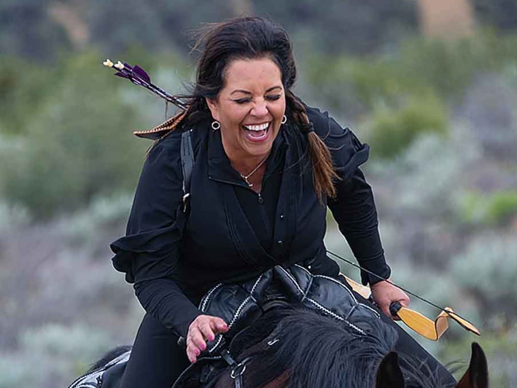 archer leaning forward laughing on horseback with blurred greenery in the background
