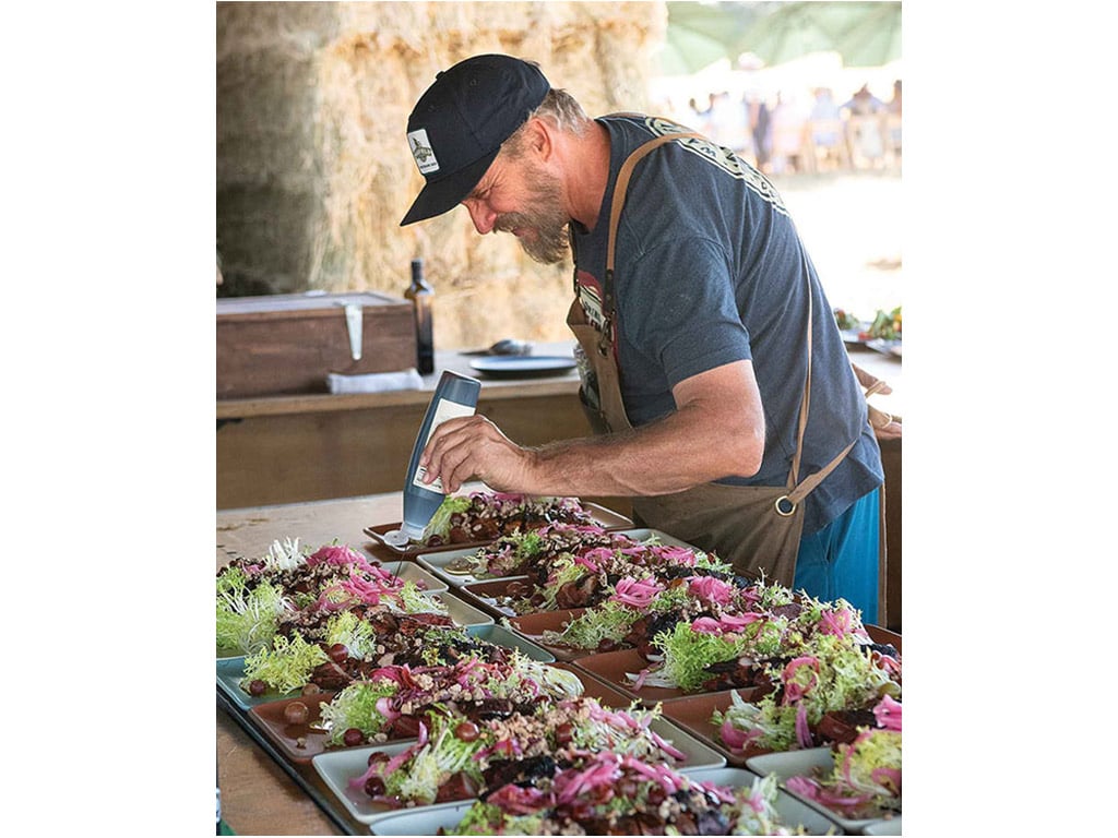 smiling chef applying ingredients to one of several plates on a table outdoors