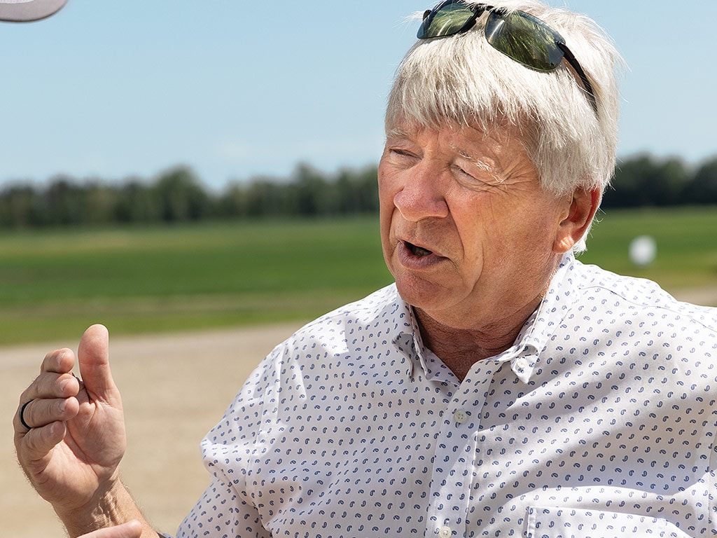 A person gesturing while speaking with blurred field and trees in the background