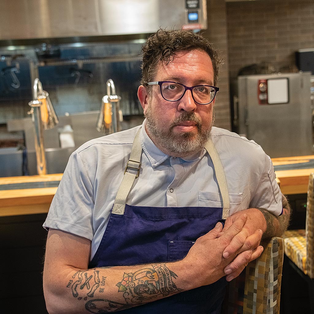 restaurant chef in apron with fingers interlocked and arm tattoos