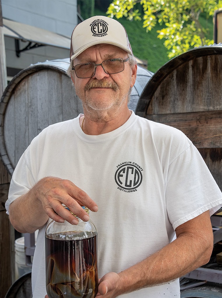 man with baseball cap and glasses holding moonshine bottle and smiling