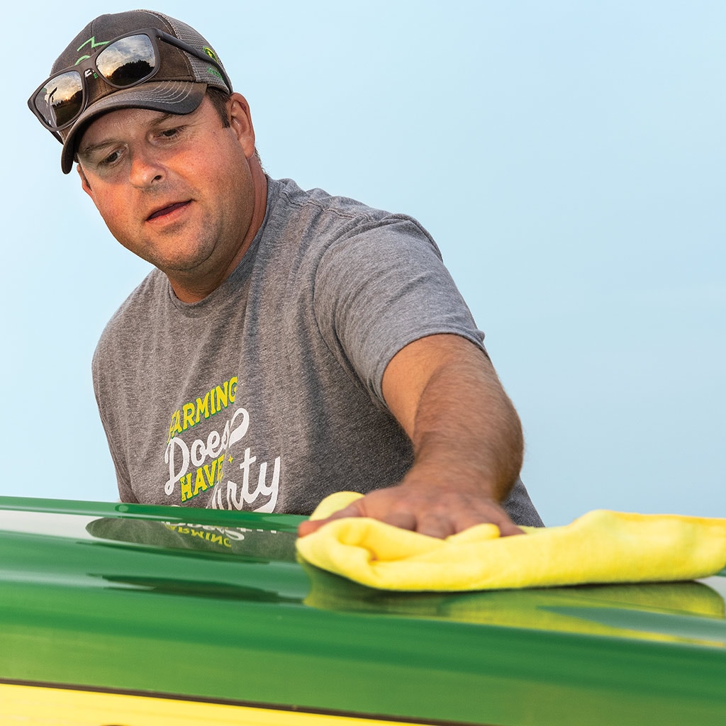 man with baseball hat polishing green Deere equipment with yellow towel