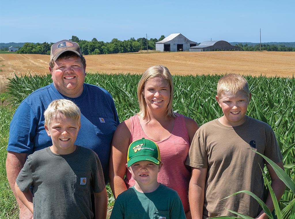 family of farmers standing in deep green field with barn in the distance