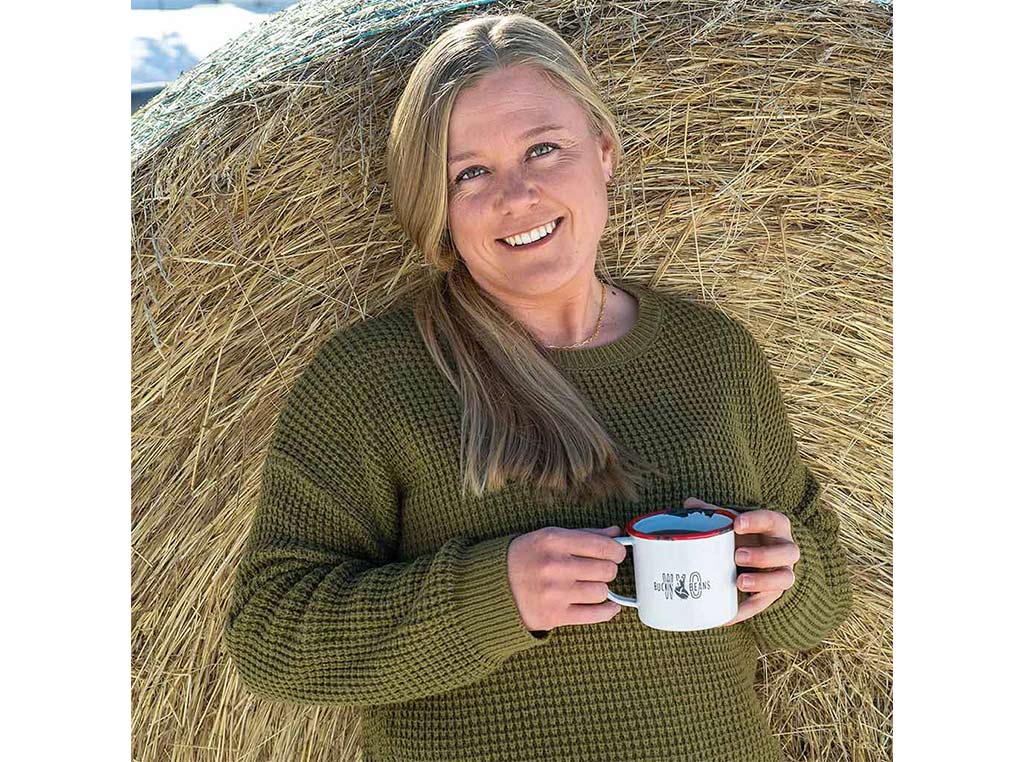 young blonde woman with olive green woven sweater and coffee mug smiling in front of hay bale