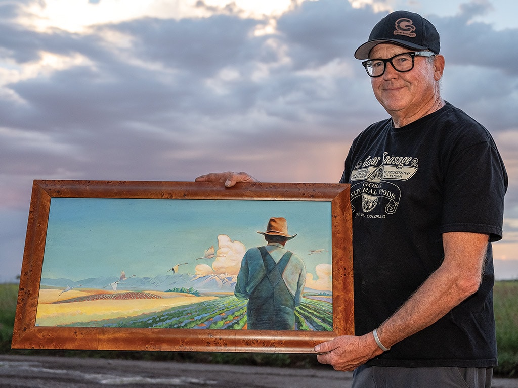 Person with black tshirt, baseball cap, and glasses presenting painting of farmer facing field of crops from behind