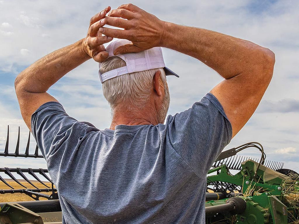Person facing away with white baseball cap and blue tshirt with hands resting on their head and John Deere equipment in the background