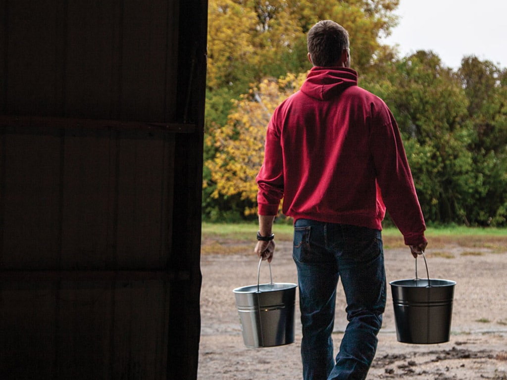 Person with red hooded sweater and jeans on walking with a metal pail in each hand with trees in the background