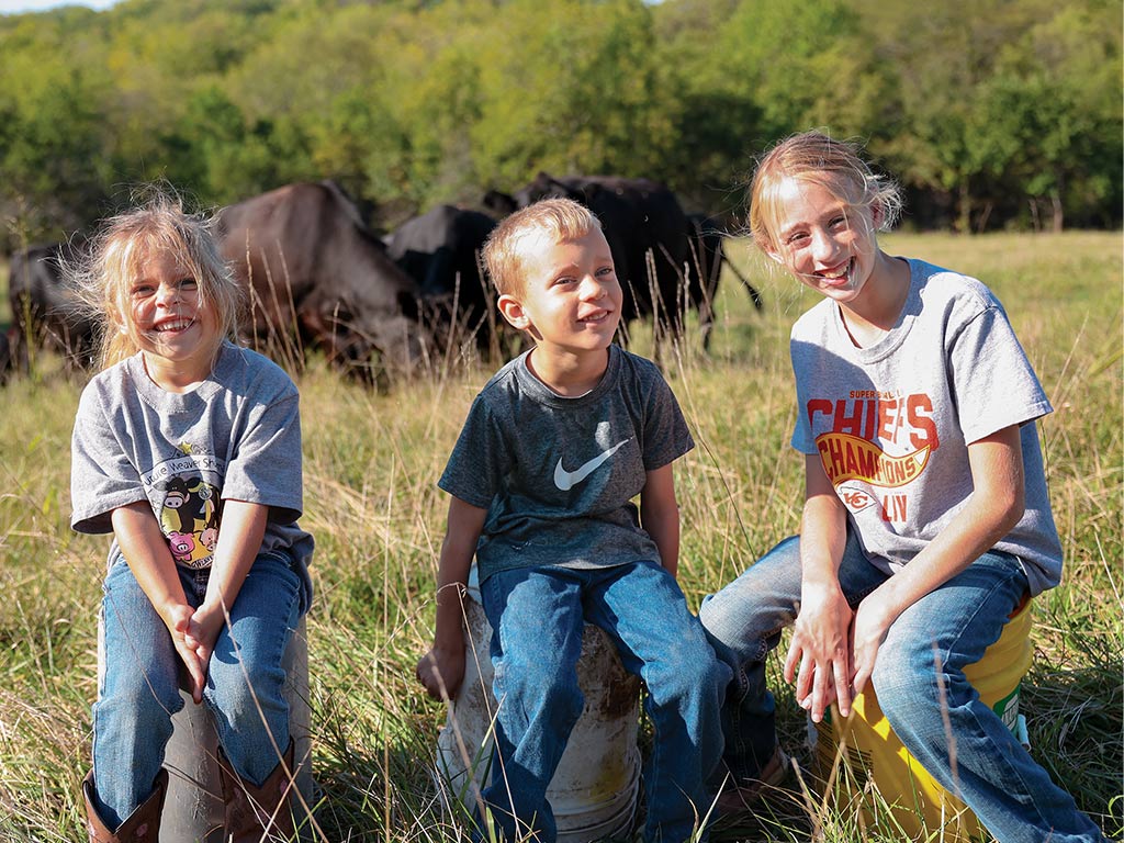 three smiling children sitting on buckets in a field with cattle