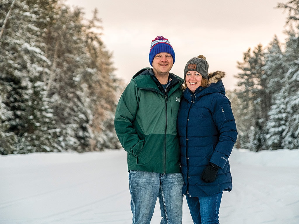 a couple in winter clothing standing in snow between two rows of evergreen trees