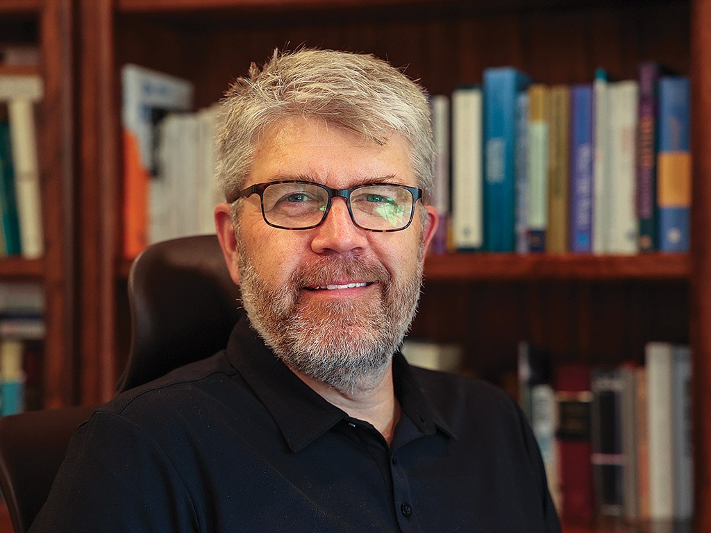 person with light hair and beard smiling with glasses and black collared shirt on in front of a bookcase