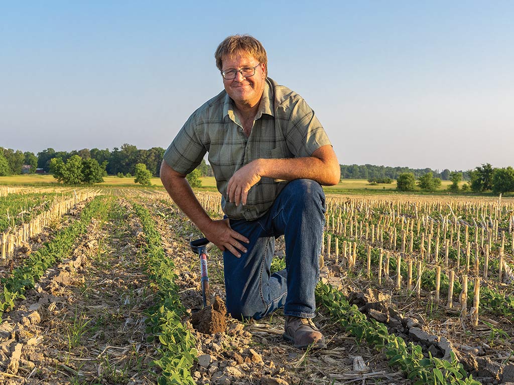 Farmer kneeling in soybean field with glasses on and shovel on the ground to their side