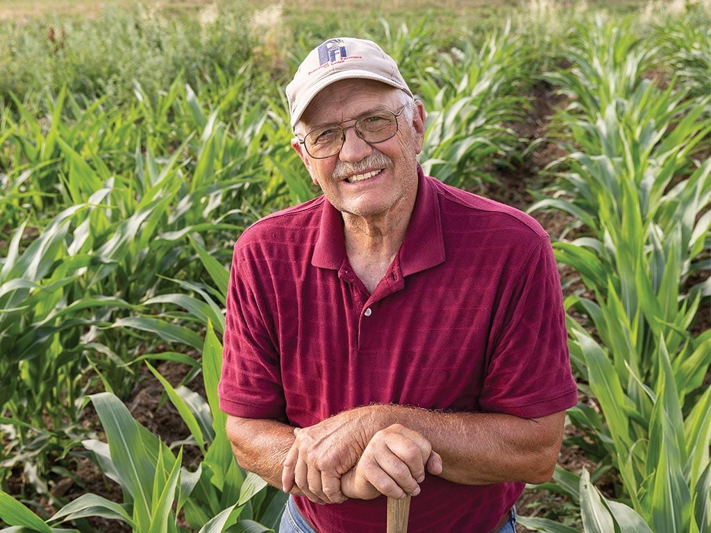 Farmer smiling in field with baseball hat and glasses resting hands on shovel handle