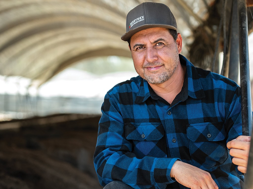 Person smiling with baseball cap and black and blue plaid flannel shirt crouched in compost tent