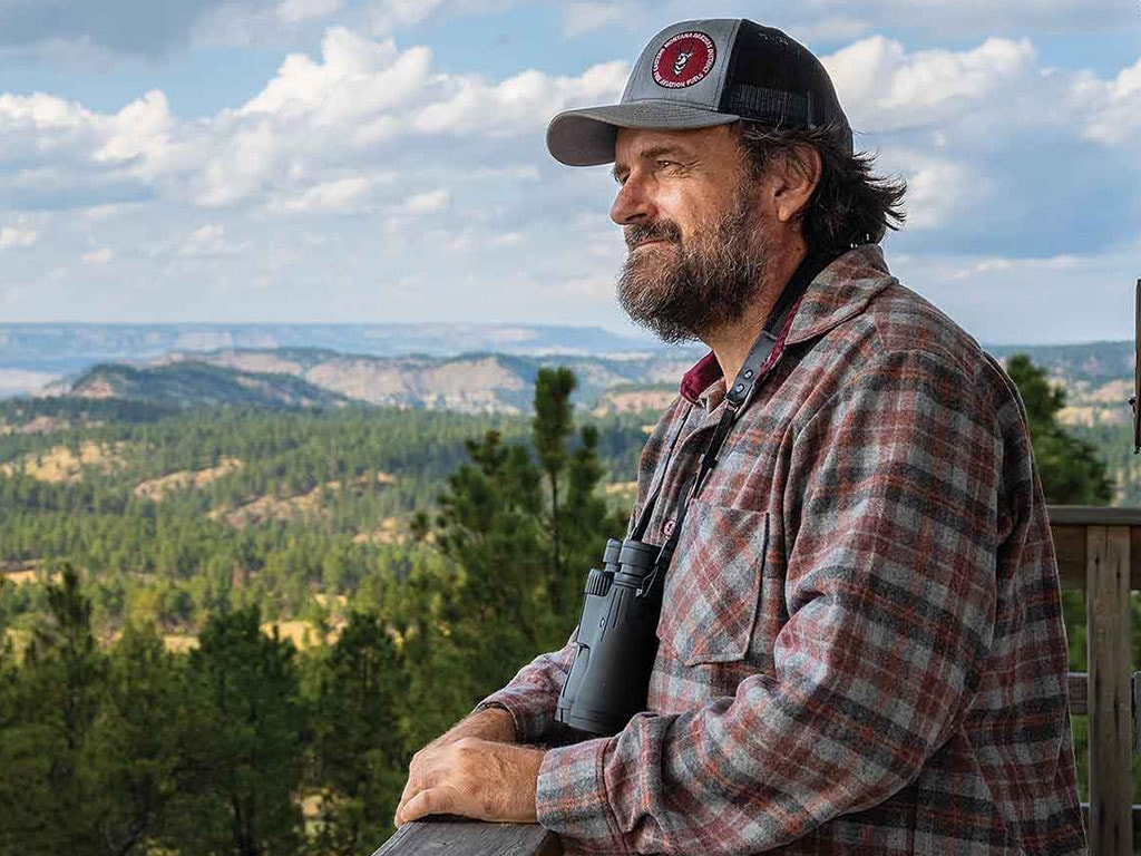 a person in a baseball cap smiling with mountains and trees in the distant background