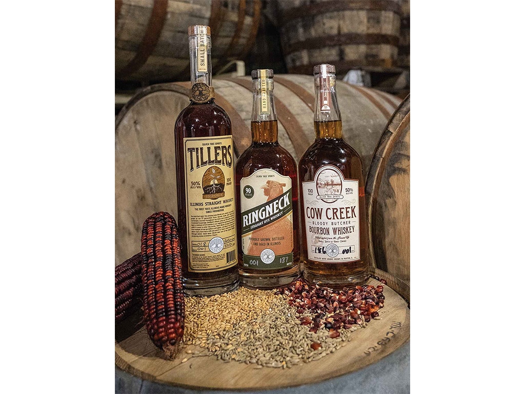 three bottles of whiskey on a wooden barrel surrounded by malt grain and red corn kernels and ears