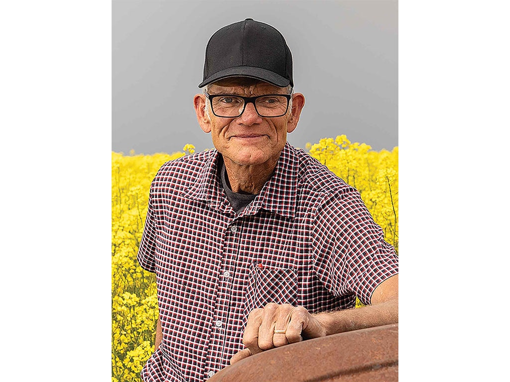 a farmer in a black baseball cap and glasses smiling in front of a bright yellow field of canola
