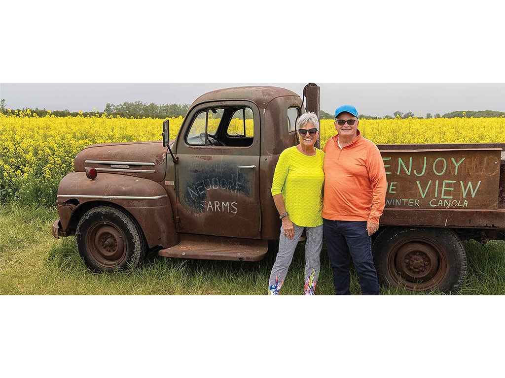 two smiling farmers in front of a rusted truck surrounded by a bright yellow canola field